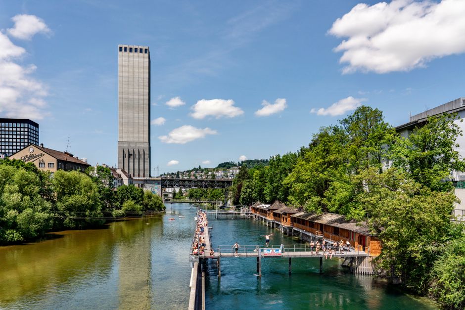 People enjoying a sunny day swimming near a city river with lush greenery and modern architecture.