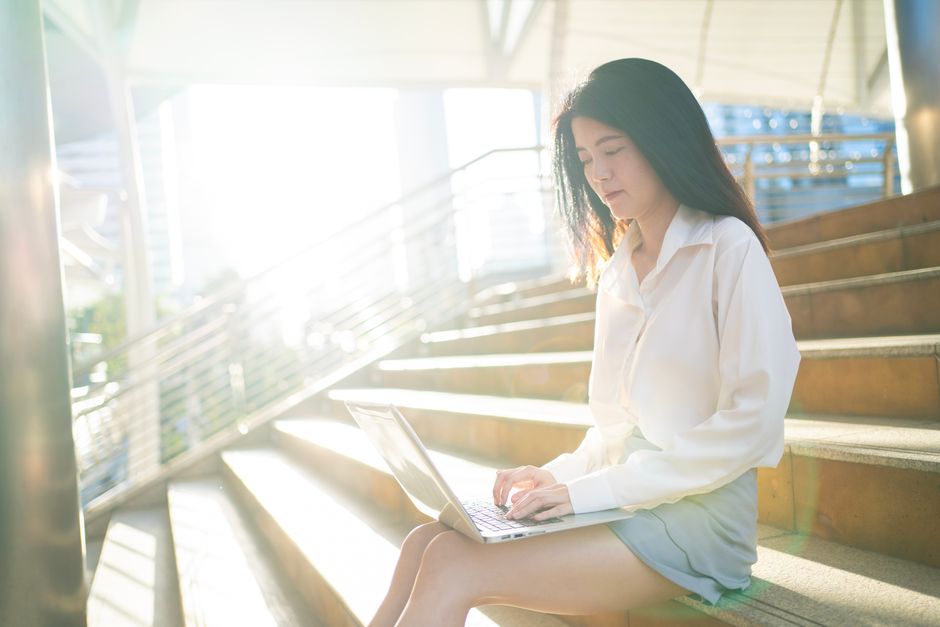 Young Asian woman working remotely on stairs with laptop, sunny urban setting in Bangkok.