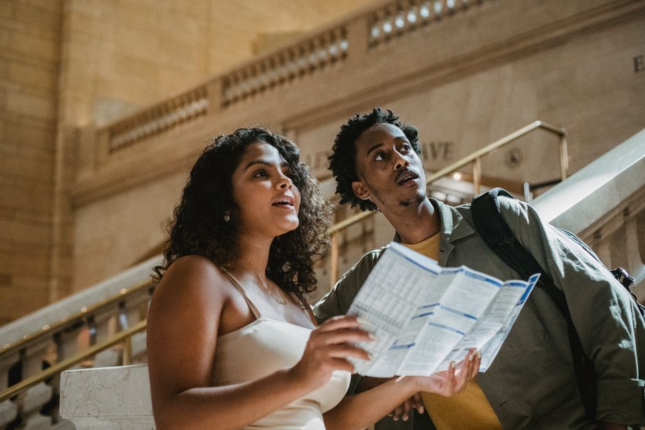 Low angle of astonished young multiracial couple in casual clothes looking away with curiosity while reading map in railway station terminal